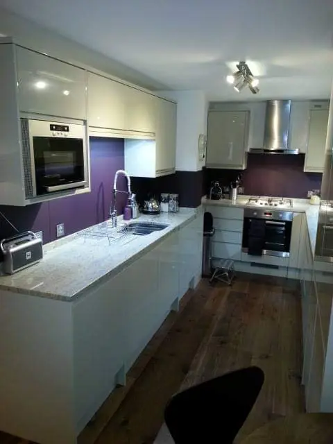 Galley kitchen with white gloss cabinets, granite counters and a purple backsplash